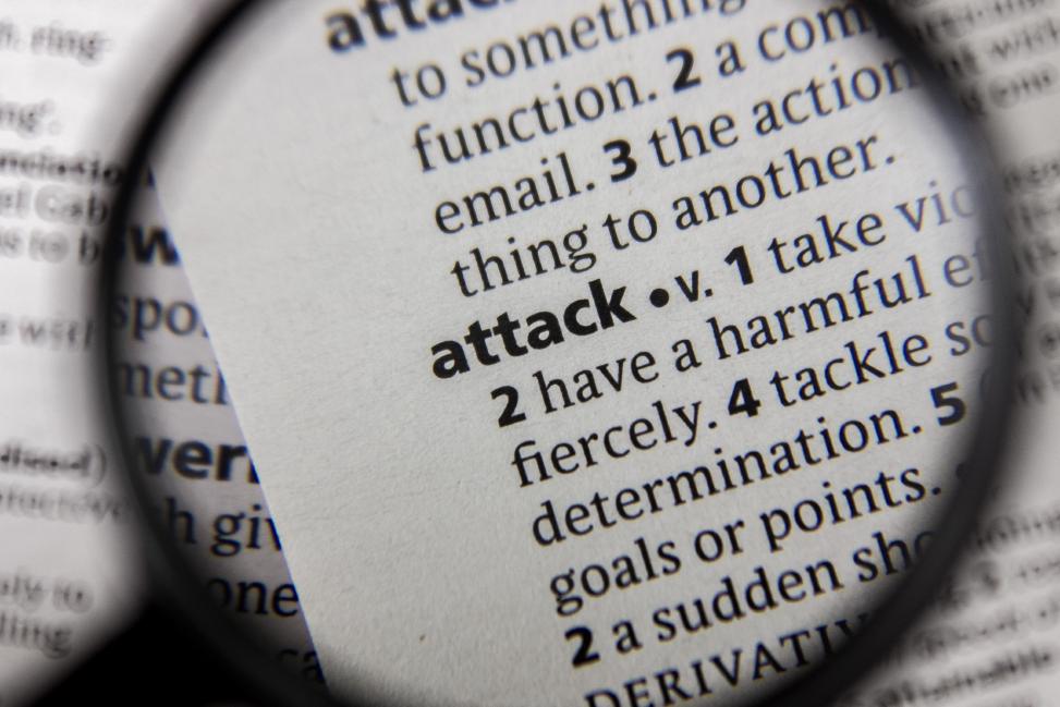 In a dictionary, under the initial letter A, the term attack, which symbolizes cyber-attacks, is shown enlarged by a magnifying glass.