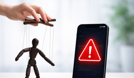 A marionette made of dark wood, representing social engineering and moved by a hand, next to a smartphone with a warning message on the display, symbolizing an attack on a two-factor authentication system. 