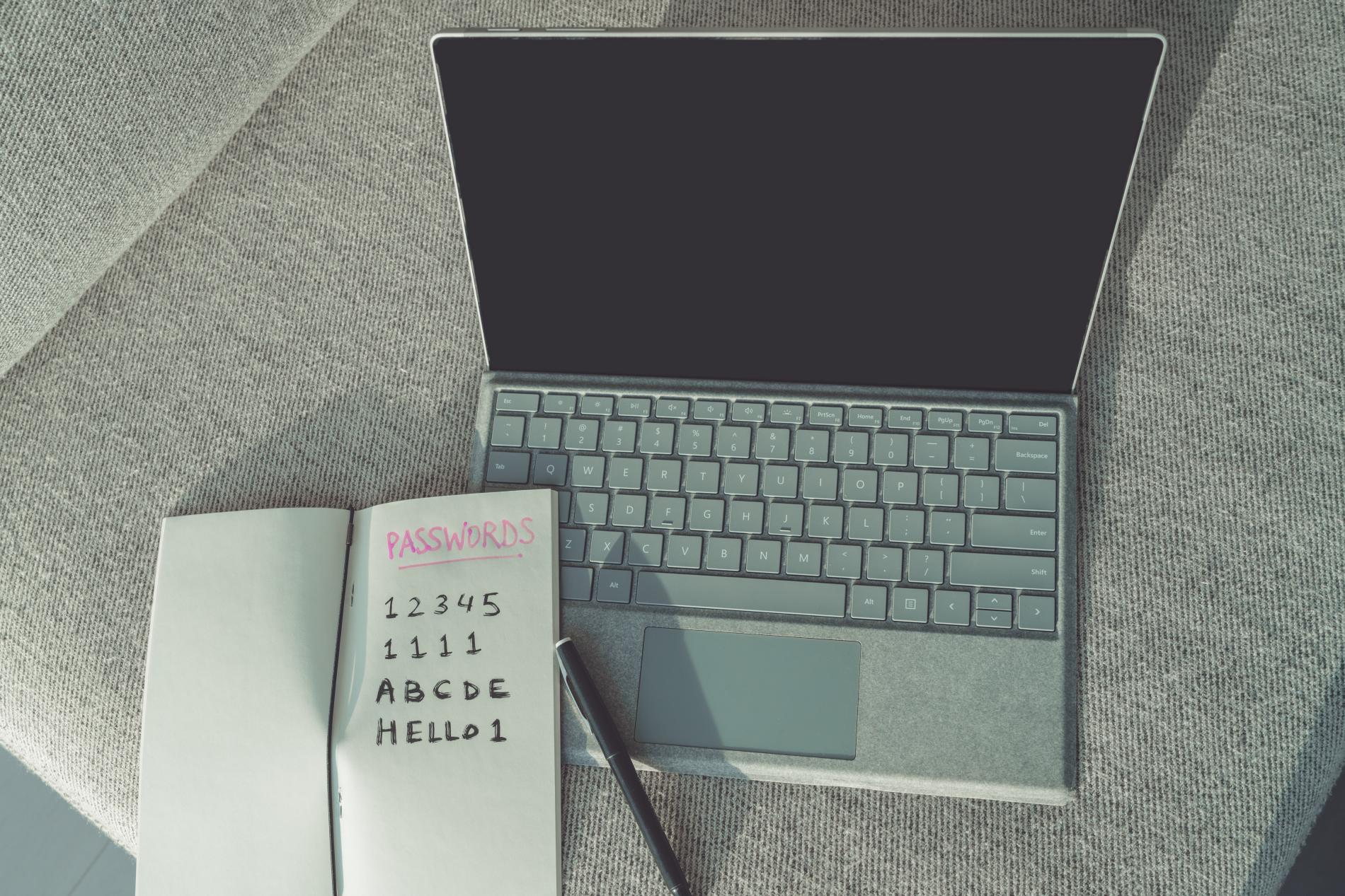 Mehrere einfache Passwörter, die in einem Notizbuch aufgeschrieben sind, liegen oben auf einem Laptop und zeigen, wie ein sicheres Passwort nicht aussieht.