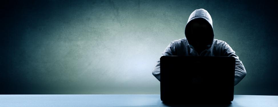 Attacking hacker sitting in front of a notebook in the darkness and wearing a hooded sweatshirt, trying to crack passwords from a user database.