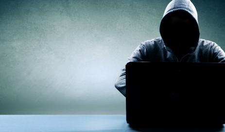 Attacking hacker sitting in front of a notebook in the darkness and wearing a hooded sweatshirt, trying to crack passwords from a user database.