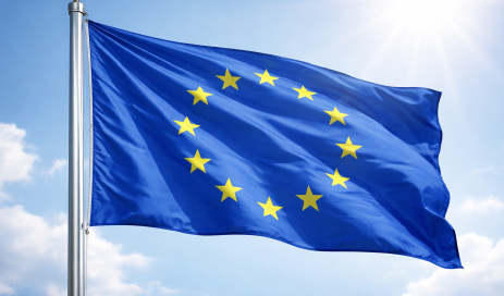 The Blue Flag of the European Union with its twelve golden stars moving in the wind on a flagpole, with the sun shining in the background.
